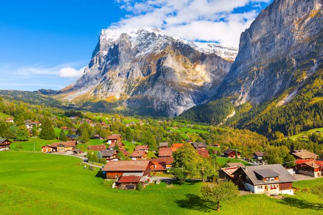Photo of aerial village view and autumn Swiss Alps mountains panorama landscape, wooden chalets on green fields and high peaks in background.