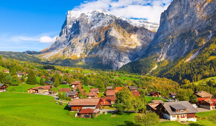Photo of aerial village view and autumn Swiss Alps mountains panorama landscape, wooden chalets on green fields and high peaks in background.