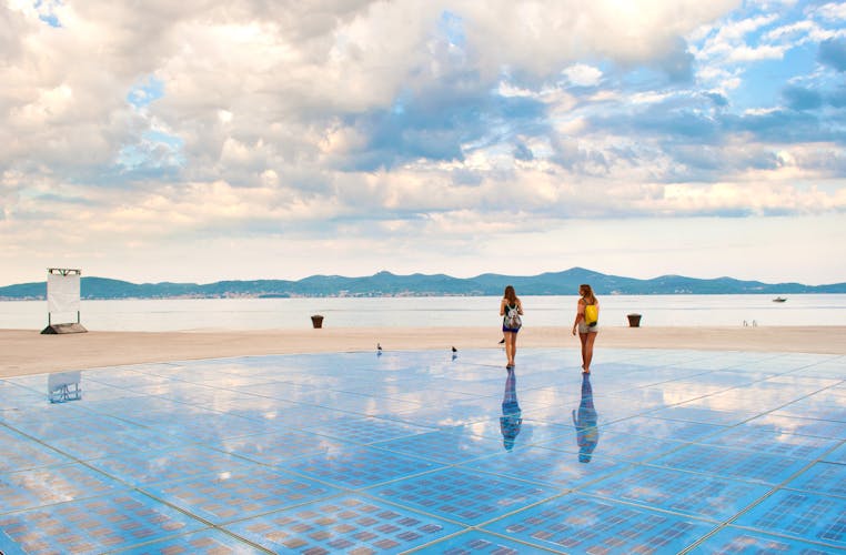 Photo of Two girls walking on top of Sun Salutation installation near the sea shore against the background of a hill range and cloudy morning sky. Zadar