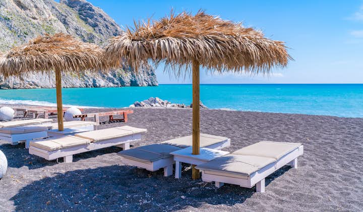 Photo of empty deck chairs and umbrellas on the black sand beach in Perissa, Santorini, Greece.