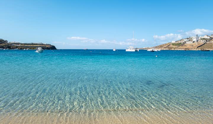 Photo of Ornos sandy tropical beach transparent sea water clear ,blue sky and moored sailboats in background.