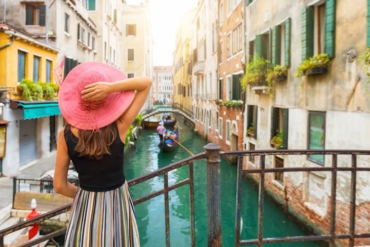 Elegant woman with red sunhat enjoys the view to a canal with passing by gondola in Venice, Italy.