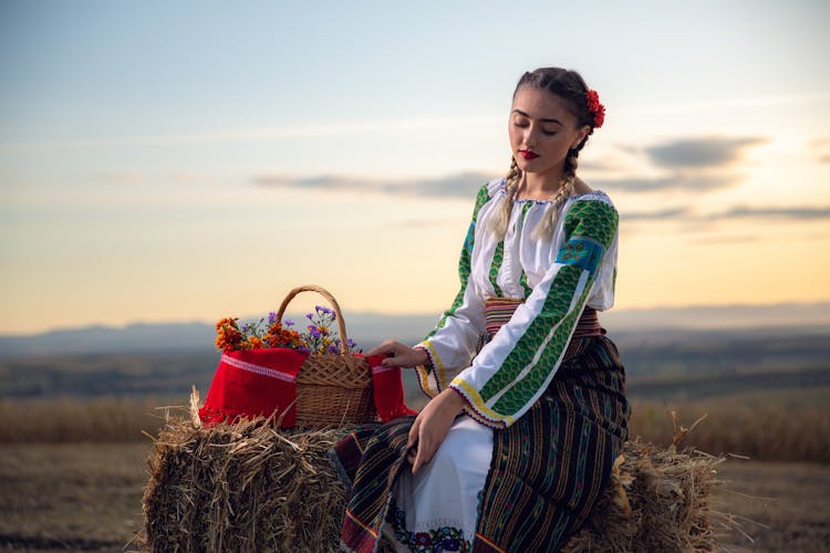  A young female in a traditional Romanian outfit with a basket of flowers on a field at sunset.