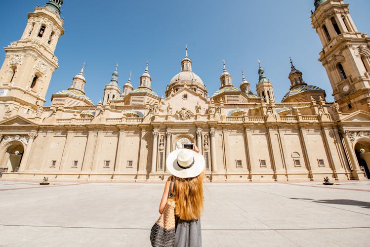 Photo of  tourist in sunhat standing back in front of the famous cathedral on the central square during the sunny weather in Zaragoza city, Spain.