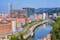 Photo of aerial view of Bilbao, Spain city downtown with a Nevion River, Zubizuri Bridge and promenade. Mountain at the background, with clear blue sky.