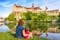  PHOTO OF VIEW OFFamily sit near medieval Sigmaringen Castle, Germany. Young woman and her baby are by river in summer. Scenic view of old famous castle on cliff on sunny day. Travel and vacation in Baden-