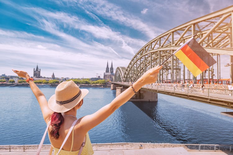 Photo of happy tourist girl holding a German flag against the Rhine bridge and Cologne Dom Cathedral. 