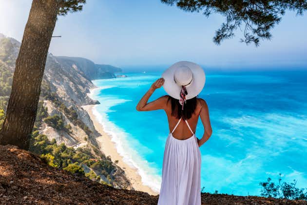 PHOTO OF A woman in a white summer dress stands on a viewing point over the turquoise, fluorescent blue sea of Egremni Beach at the island of Lefkada, Greece .