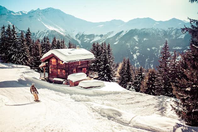 photo of landscape view of a traditional Swiss house with the roof covered by snow and mountains in the background and a snowboarder on the left , in the town of "Verbier" in Switzerland.