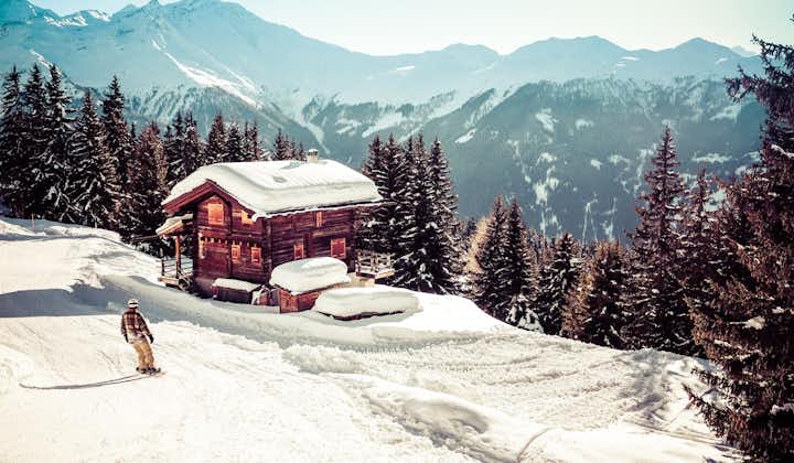 photo of landscape view of a traditional Swiss house with the roof covered by snow and mountains in the background and a snowboarder on the left , in the town of "Verbier" in Switzerland.