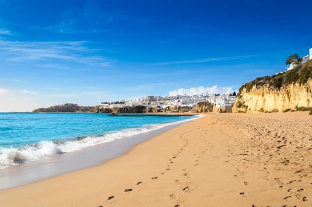 Photo of waves crashing against wide sandy beach Praia de Albufeira and view over cliffs and white houses in the old town, Portugal.